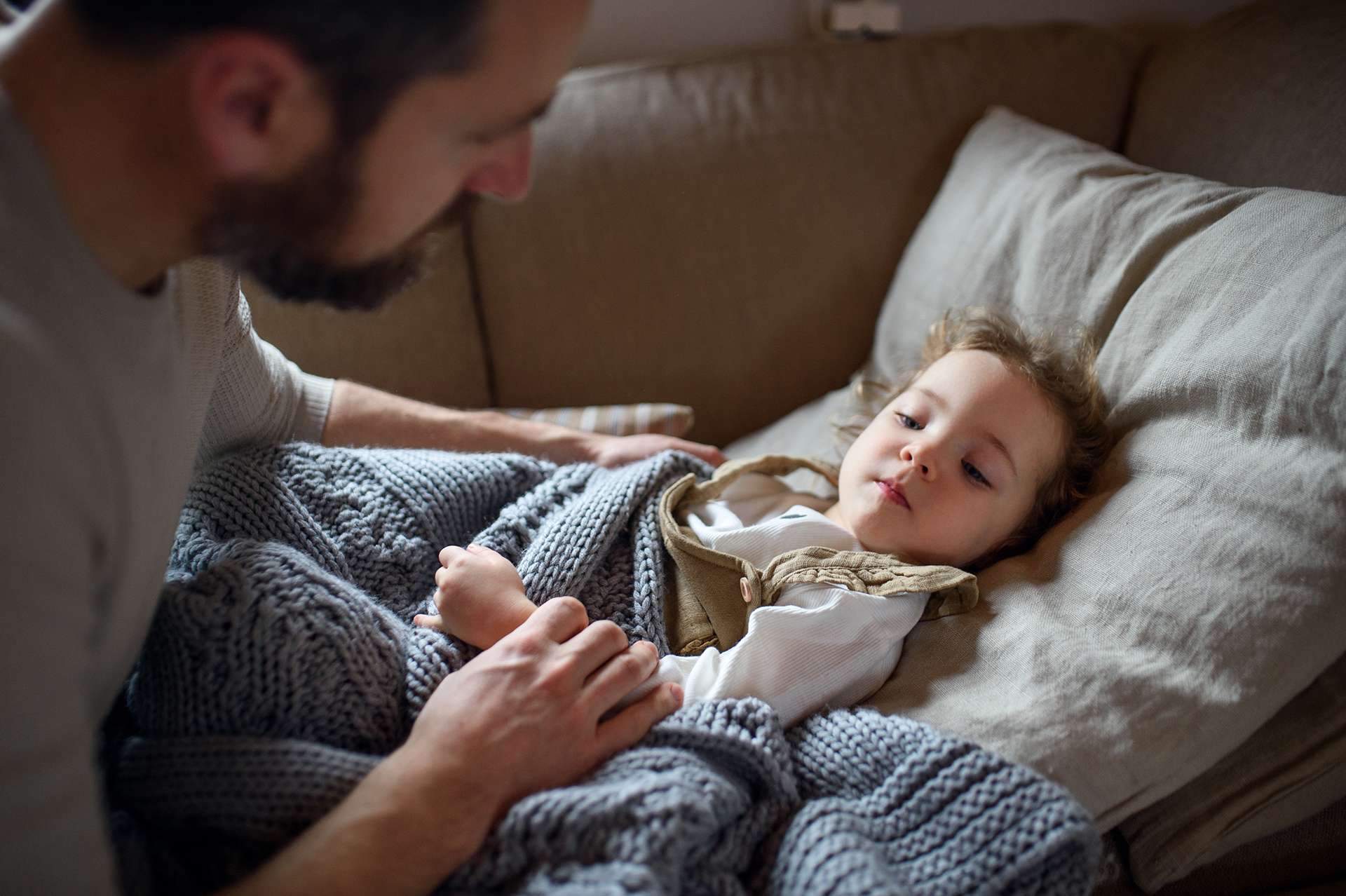 A child lies on the couch and suffers from an ear infection, and the father sits next to the child taking care of the child.